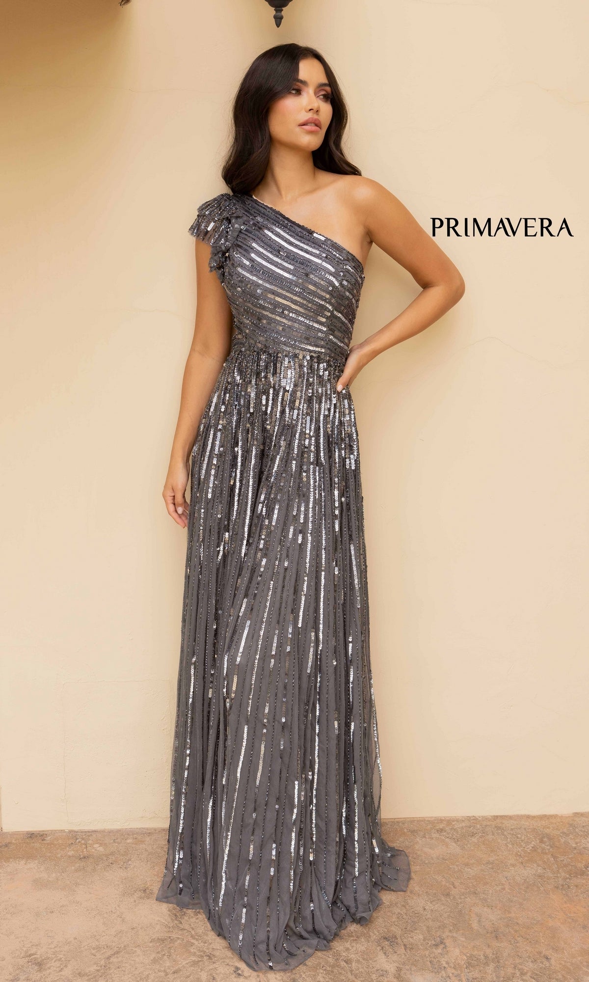 A woman in the Strappy-Back One-Shoulder Long Sequin Formal Dress, a floor-length silver gown, poses against a beige wall with